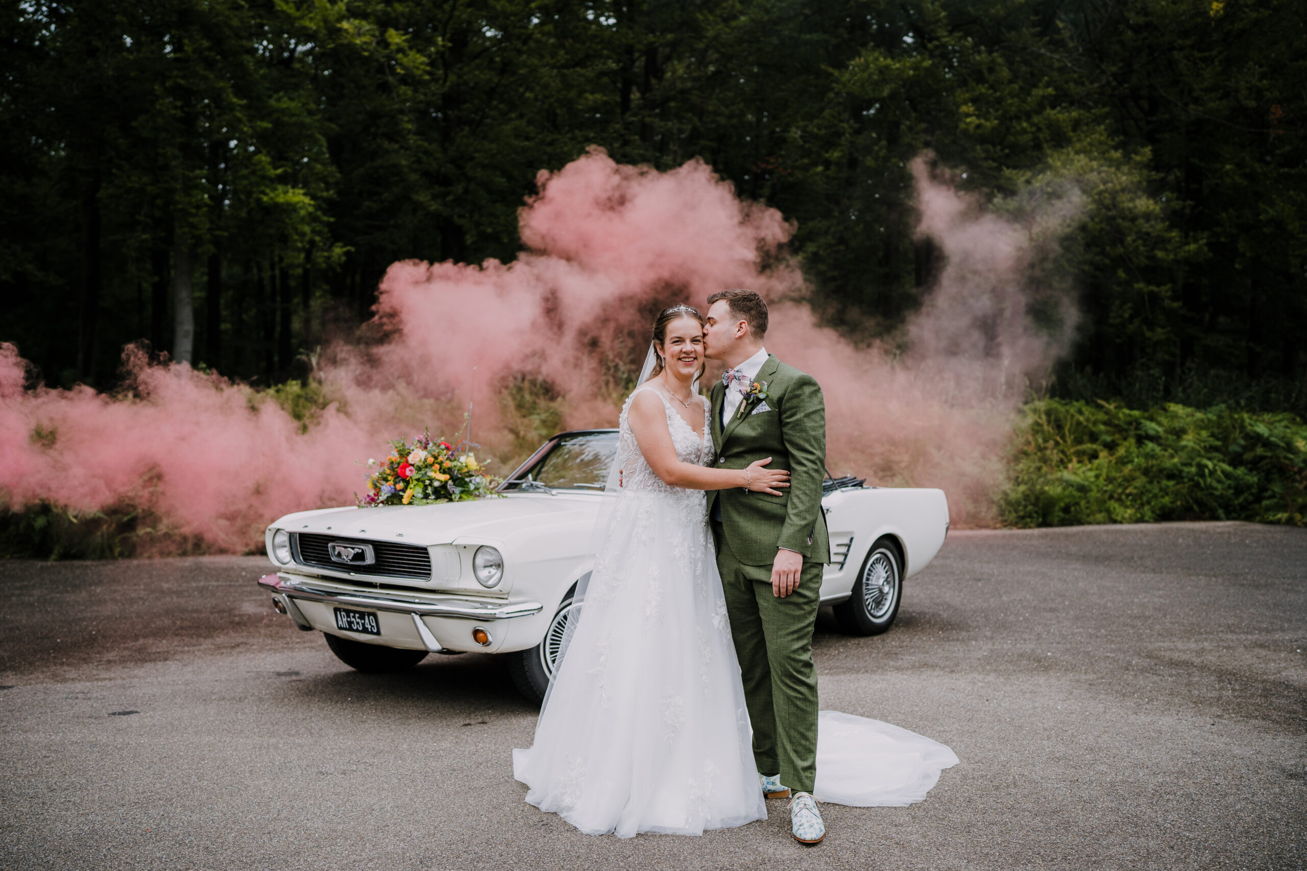 Een bruidspaar dat voor een Ford Mustang op de parkeerplaats bij Landgoed Staverden poseert, met roze rook op de achtergrond.